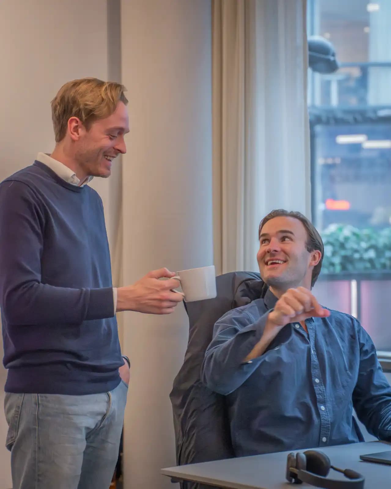 Two colleagues in an office. One is standing with a coffee cup, the other is sitting at a desk and laughing.